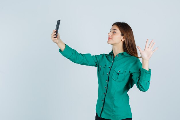 Jeune fille en chemisier vert, pantalon noir prenant selfie avec téléphone, levant la main pour dire bonjour et à la joyeuse vue de face.