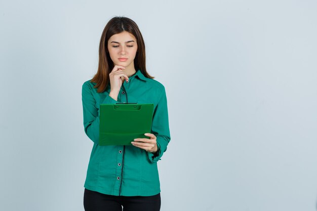 Jeune fille en chemisier vert, pantalon noir regardant le presse-papiers, soutenant le menton et regardant focalisé, vue de face.