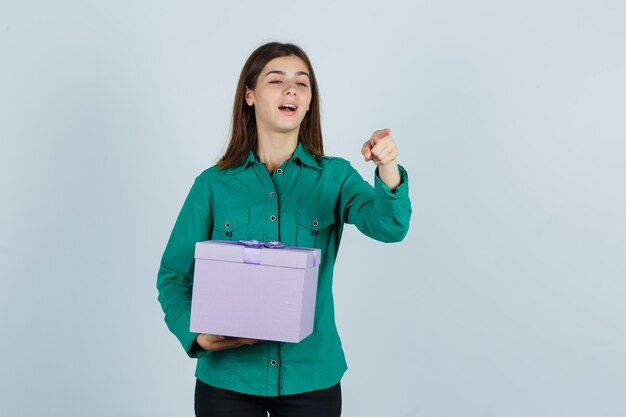 Jeune fille en chemisier vert, pantalon noir tenant une boîte-cadeau, pointant la caméra avec l'index et à la recherche focalisée, vue de face.