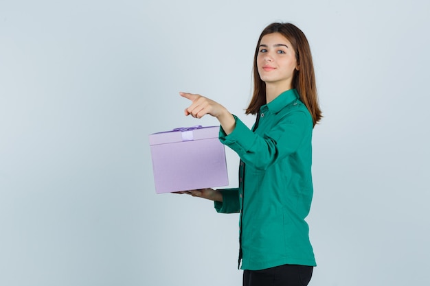 Jeune fille en chemisier vert, pantalon noir tenant la boîte-cadeau, pointant vers l'extérieur avec l'index et à la joyeuse, vue de face.