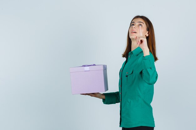 Jeune fille en chemisier vert, pantalon noir tenant la boîte-cadeau, pointant vers le haut avec l'index et à la vue de face, focalisée.