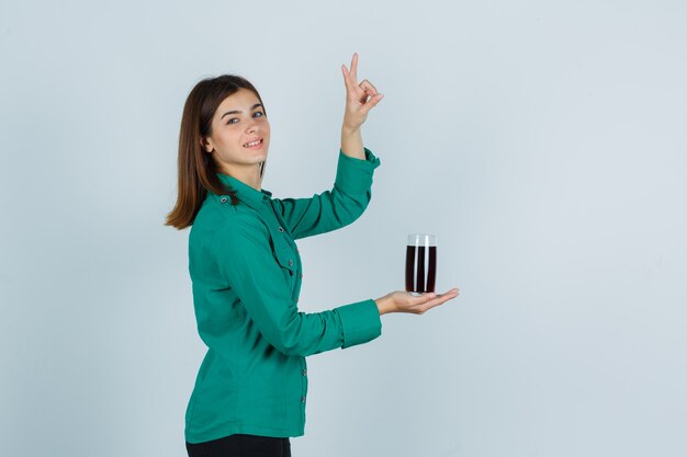 Jeune fille en chemisier vert, pantalon noir tenant un verre de liquide noir, montrant le geste de paix et l'air heureux, vue de face.