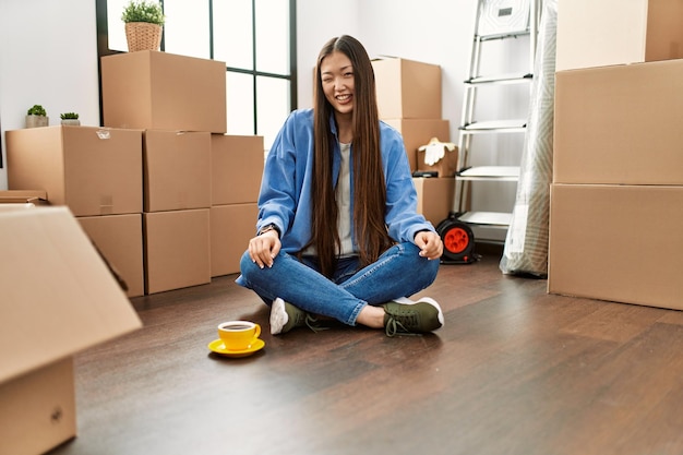 Jeune fille chinoise assise sur le sol dans une nouvelle maison en faisant un clin d'œil en regardant la caméra avec une expression sexy visage joyeux et heureux