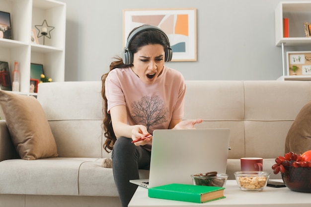 Jeune fille en colère portant des écouteurs tenant un stylo utilisé un ordinateur portable assis sur un canapé derrière une table basse dans le salon
