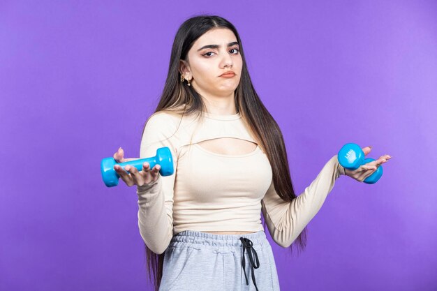 Jeune fille confuse tenant deux haltères et regardant la caméra Une jeune fille en forme se tient sur fond violet Photo de haute qualité