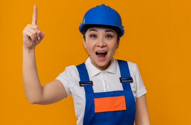 Jeune fille de constructeur asiatique surprise avec un casque de sécurité bleu pointant vers le haut