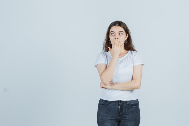 Jeune fille couvrant la bouche avec la main, regardant de côté en t-shirt, jeans et à la surprise. vue de face.