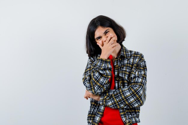 Jeune fille couvrant la bouche avec la main, riant en chemise à carreaux et t-shirt rouge et l'air heureux, vue de face.