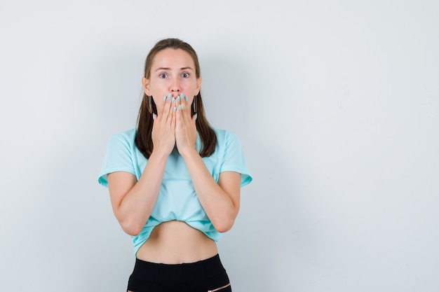 Jeune fille couvrant la bouche avec des paumes en t-shirt turquoise, un pantalon et l'air effrayé, vue de face.