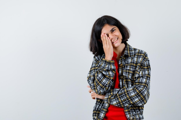 Jeune fille couvrant les yeux avec la main, riant en chemise à carreaux et t-shirt rouge et l'air heureux, vue de face.