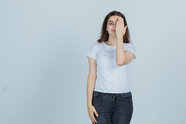 Jeune fille couvrant les yeux avec la main en t-shirt, jeans et regardant heureux, vue de face.