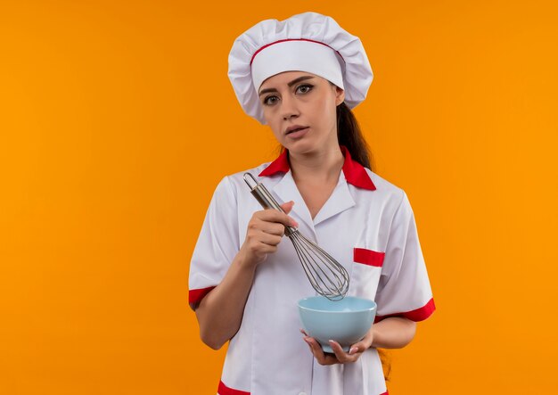 Jeune fille de cuisinier caucasien agacé en uniforme de chef détient bol et fouet isolé sur fond orange avec copie espace