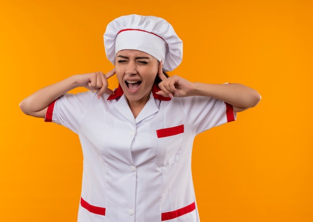 Photo gratuite jeune fille de cuisinier caucasien agacé en uniforme de chef ferme les oreilles avec les doigts isolés sur fond orange avec espace de copie