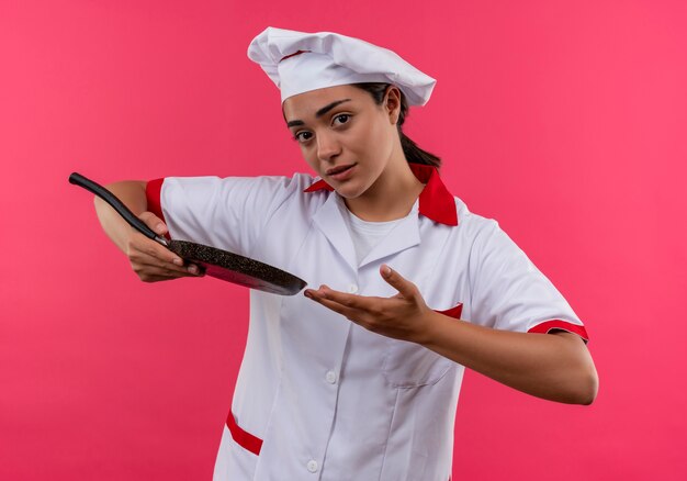 Jeune fille de cuisinier caucasien anxieux en uniforme de chef détient une poêle et des points avec la main isolé sur fond rose avec copie espace