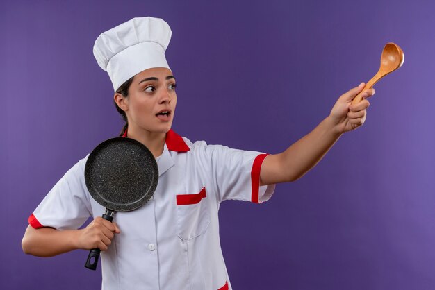 Jeune fille de cuisinier caucasien anxieux en uniforme de chef tient une poêle et pointe sur le côté avec une cuillère en bois isolé sur fond violet avec espace copie