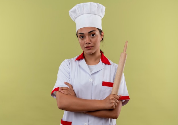 Jeune fille de cuisinier caucasien confiant en uniforme de chef croise les bras et tient le rouleau à pâtisserie isolé sur fond vert avec espace copie