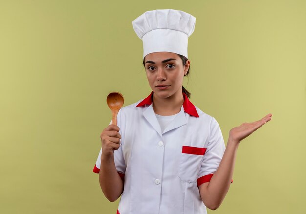 Jeune fille de cuisinier caucasien confiant en uniforme de chef détient une cuillère en bois et garde la main ouverte isolé sur mur vert avec espace copie