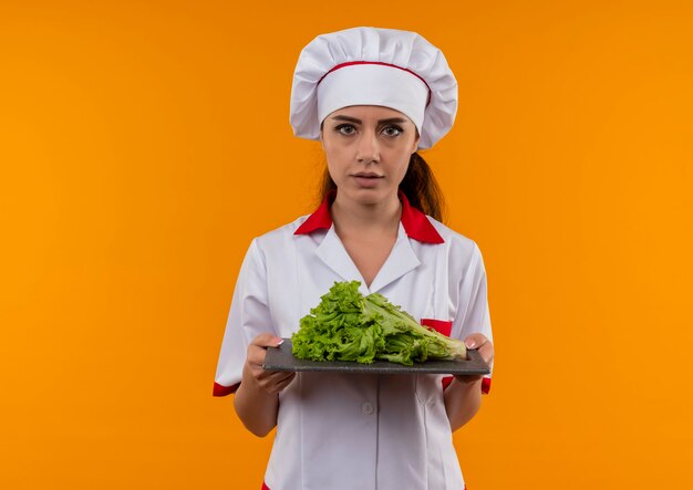Jeune fille de cuisinier caucasien confiant en uniforme de chef détient la salade sur une planche à découper isolé sur un mur orange avec copie espace