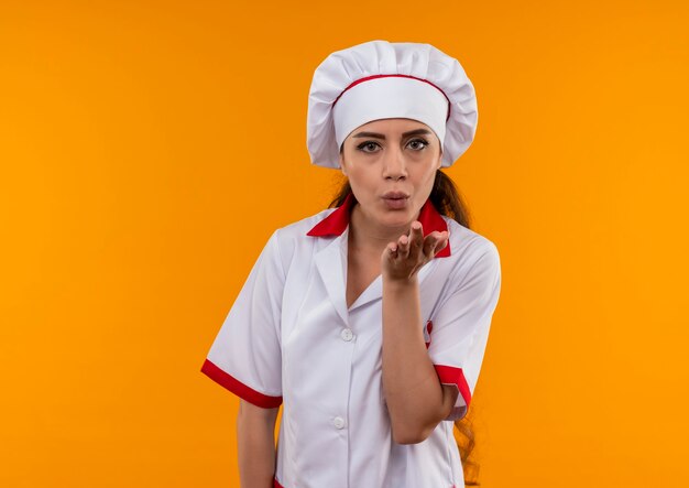 Jeune fille de cuisinier caucasien confiant en uniforme de chef envoie baiser avec la main isolée sur un mur orange avec espace copie