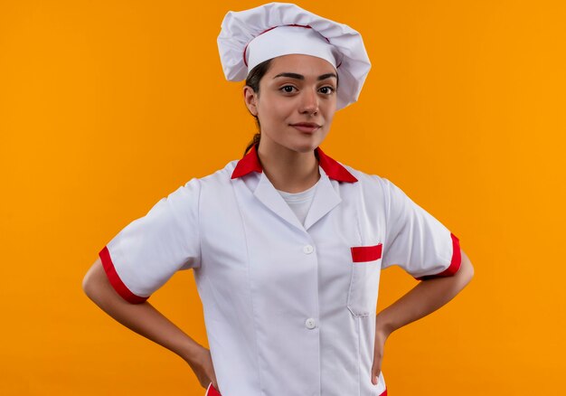 Jeune fille de cuisinier caucasien confiant en uniforme de chef met les mains sur la taille isolé sur un mur orange avec copie espace