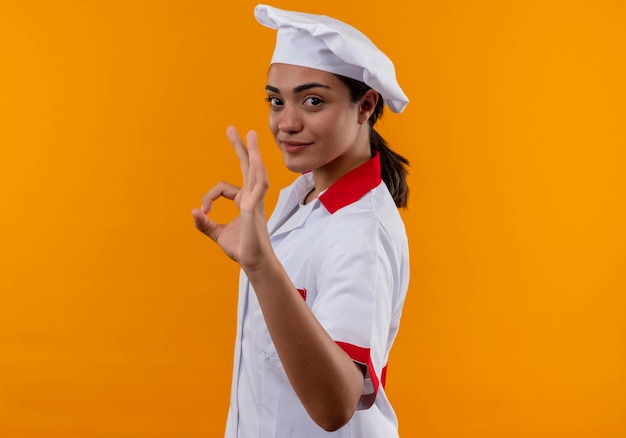 Jeune fille de cuisinier caucasien confiant en uniforme de chef se tient sur le côté et les gestes ok signe de la main isolé sur un mur orange avec copie espace