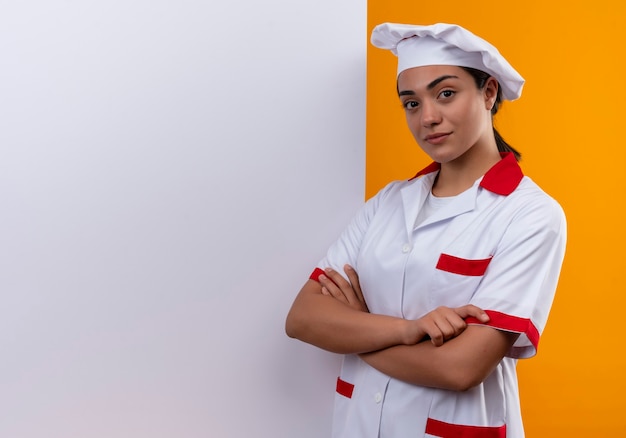 Jeune fille de cuisinier caucasien confiant en uniforme de chef se tient devant un mur blanc et croise les bras isolés sur un mur orange avec espace de copie