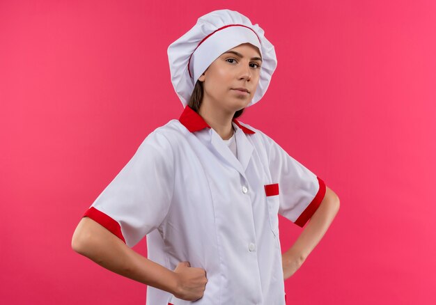 Jeune fille de cuisinier caucasien confiant en uniforme de chef tient les poings sur la taille et regarde la caméra isolée sur l'espace rose avec copie espace