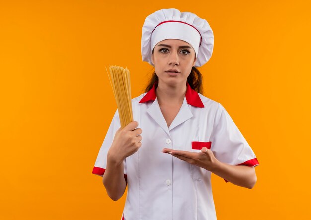 Jeune fille de cuisinier caucasien confus en uniforme de chef tient et pointe à bouquet de spaghetti isolé sur un mur orange avec copie espace