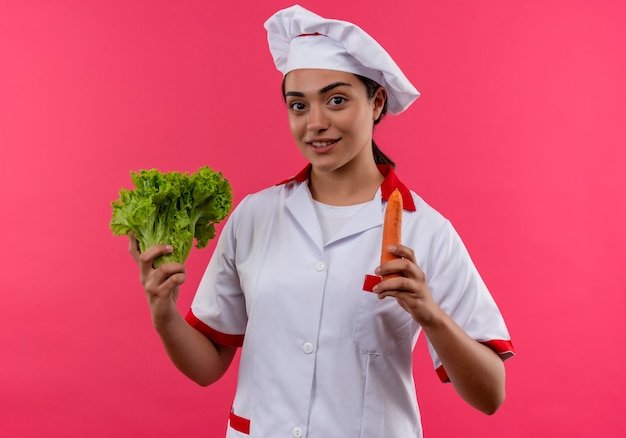 Jeune fille de cuisinier caucasien heureux en uniforme de chef détient salade et carotte isolé sur mur rose avec espace copie