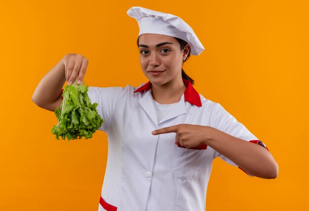 Jeune fille de cuisinier caucasien heureux en uniforme de chef détient la salade à l'envers dowm et points avec le doigt isolé sur le mur orange avec copie espace