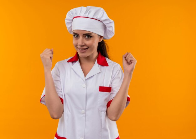 Jeune fille de cuisinier caucasien heureux en uniforme de chef lève les poings isolé sur mur orange avec espace copie