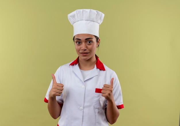 Jeune fille de cuisinier caucasien heureux en uniforme de chef pouces vers le haut des deux mains isolé sur mur vert avec espace copie