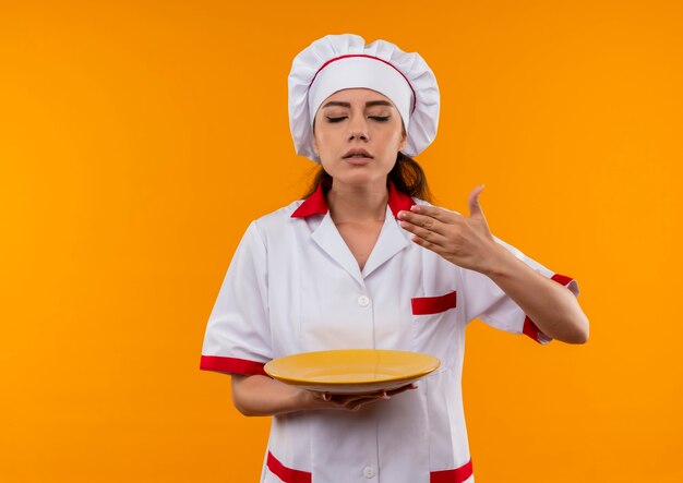 Jeune fille de cuisinier caucasien heureux en uniforme de chef tient la plaque et fait semblant de sentir isolé sur un mur orange avec copie espace