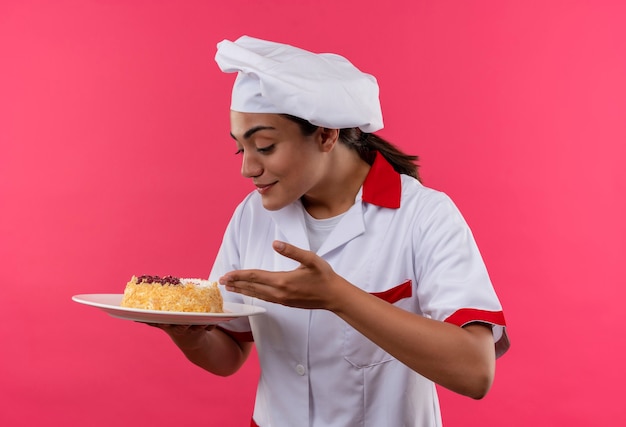 Photo gratuite jeune fille de cuisinier caucasien heureux en uniforme de chef tient et regarde le gâteau sur la plaque isolée sur le mur rose avec copie espace