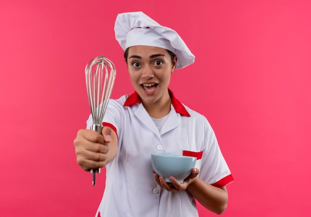 Jeune fille de cuisinier caucasien surpris en uniforme de chef détient bol et tient fouet isolé sur mur rose avec espace copie