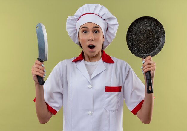 Jeune fille de cuisinier caucasien surpris en uniforme de chef détient un couteau et une poêle à frire isolé sur un espace vert avec copie espace