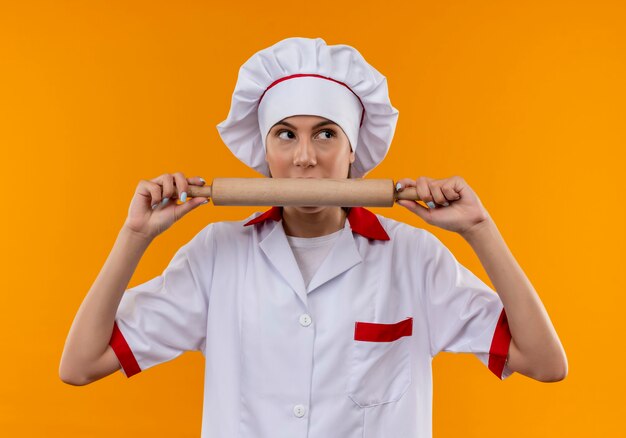 Jeune fille de cuisinier caucasien surpris en uniforme de chef détient et fait semblant de mordre le rouleau à pâtisserie isolé sur fond orange avec copie espace