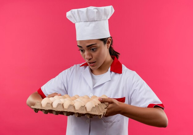 Jeune fille de cuisinier caucasien surpris en uniforme de chef détient lot d'oeufs isolé sur mur rose avec copie espace