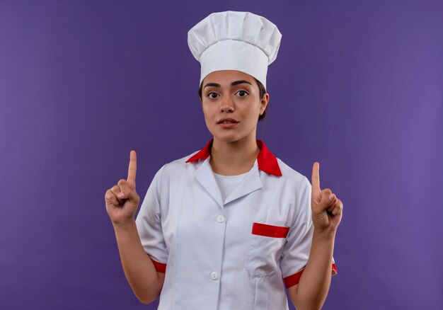 Jeune fille de cuisinier caucasien surpris en uniforme de chef pointe avec les deux mains isolé sur mur violet avec espace copie