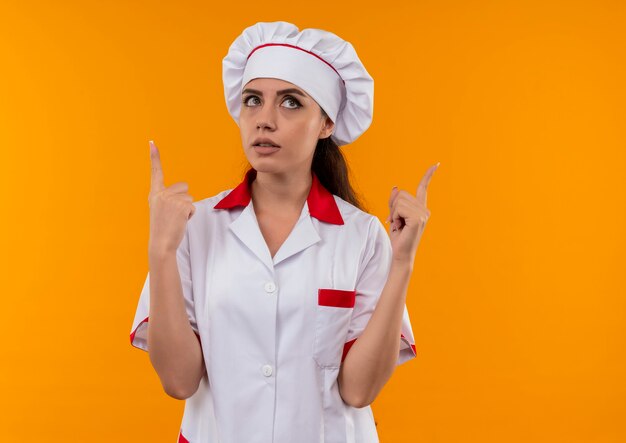 Jeune fille de cuisinier caucasien surpris en uniforme de chef regarde et pointe vers le haut isolé sur un mur orange avec copie espace