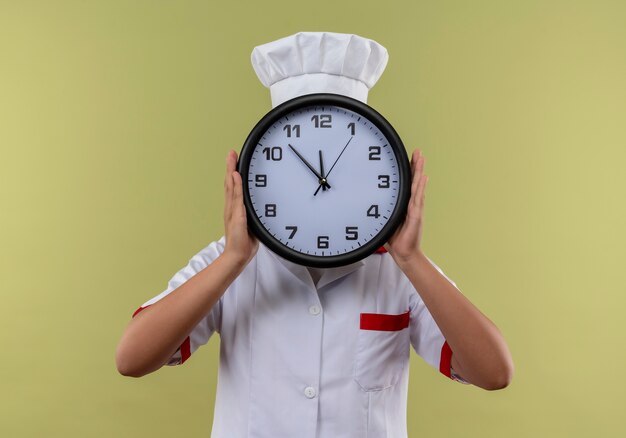 Jeune fille de cuisinier de race blanche en uniforme de chef ferme le visage avec horloge isolé sur fond vert avec espace copie