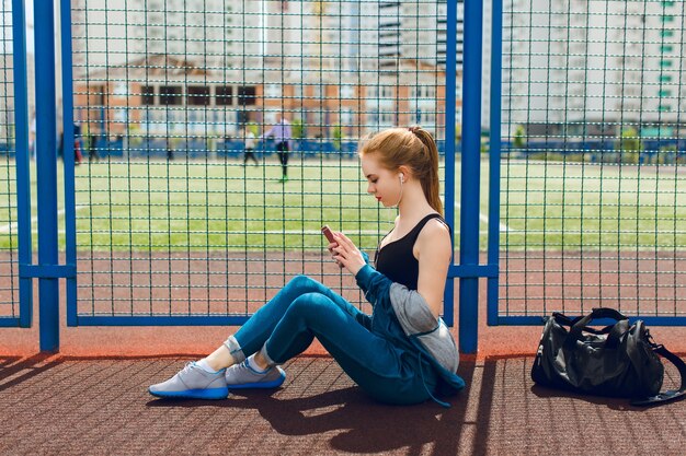 Une jeune fille dans un costume de sport bleu avec un haut noir est assise près d'une clôture sur le stade. Elle écoute la musique avec des écouteurs.