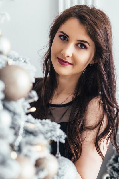 Jeune fille dans une robe noire pose devant un sapin de Noël brillant