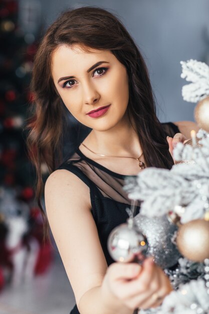 Jeune fille dans une robe noire pose devant un sapin de Noël brillant