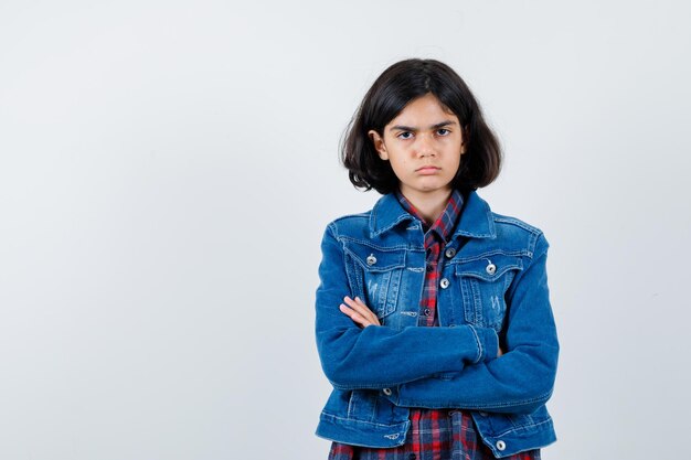 Jeune fille debout les bras croisés en chemise à carreaux et veste en jean et l'air sérieux. vue de face.