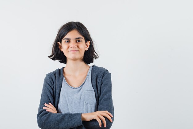 Jeune fille debout les bras croisés en t-shirt gris clair et sweat à capuche zippé gris foncé et à mignon,