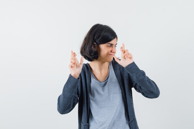 Photo gratuite jeune fille debout les doigts croisés en t-shirt gris clair et sweat à capuche zippé gris foncé et à mignon