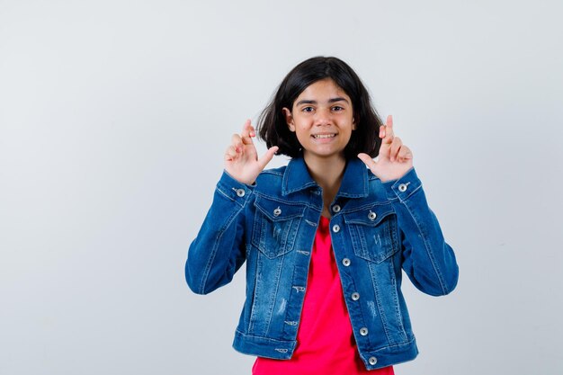 Jeune fille debout les doigts croisés en t-shirt rouge et veste en jean et l'air heureux. vue de face.