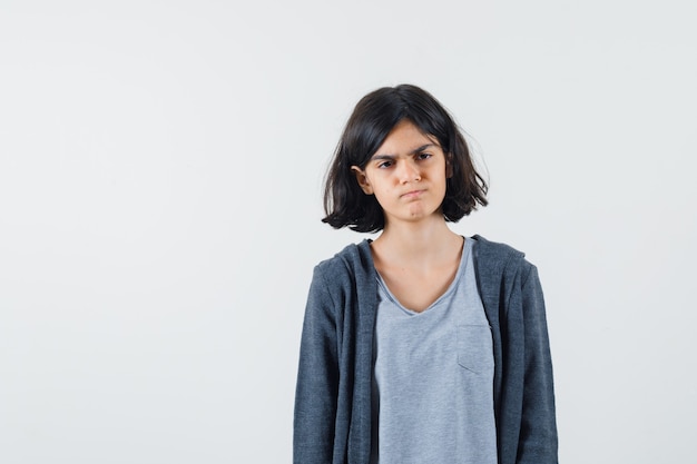 Jeune fille debout droite et posant à la caméra en t-shirt gris clair et sweat à capuche zippé gris foncé et à la recherche de sérieux.