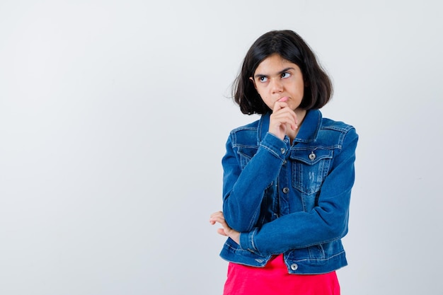 Jeune fille debout en pensant pose en t-shirt rouge et veste en jean et regardant pensive, vue de face.